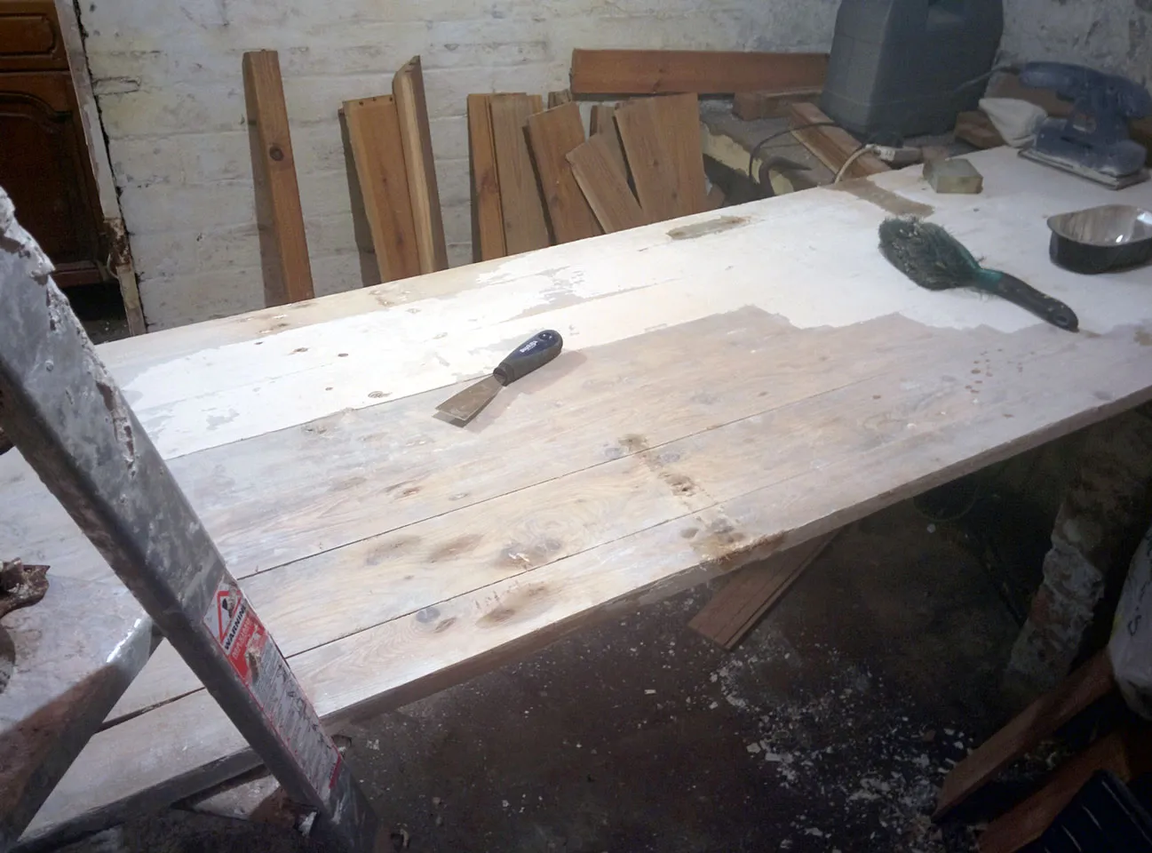 An old door laid flat in a basement with part of its white paint scraped back to bare wood, a scraper and wire brush resting on top.