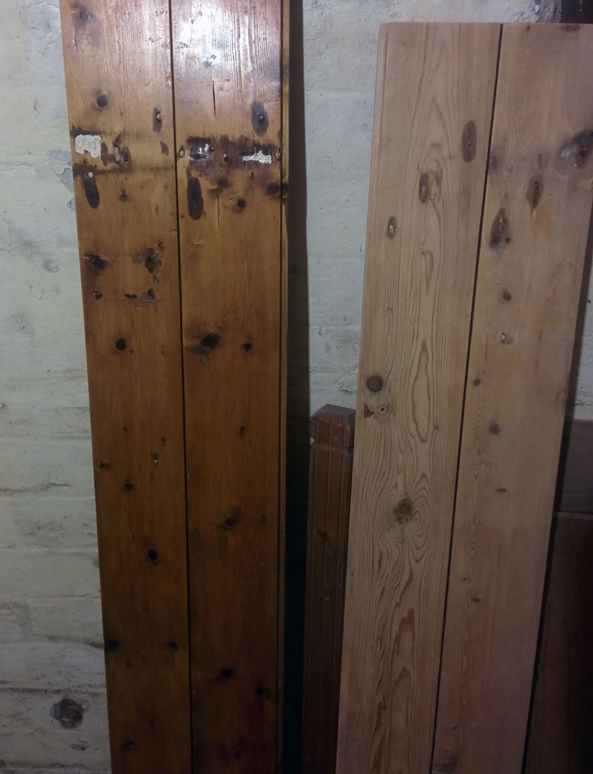 Two pairs of pine planks leaning against a whitewashed wall, the left pair darkened with oil and the right pair still pale.