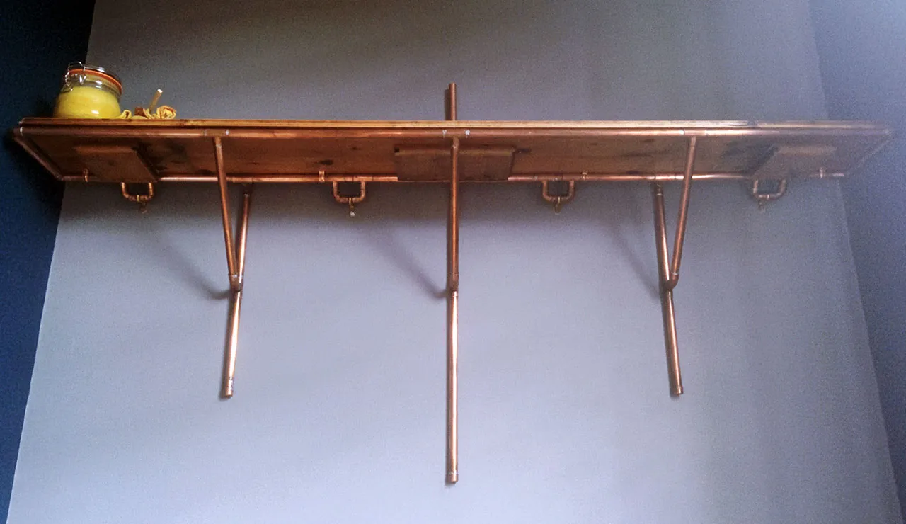 The finished wooden shelf mounted on a grey wall on three copper pipe supports, with hooks beneath and a jar on one end.