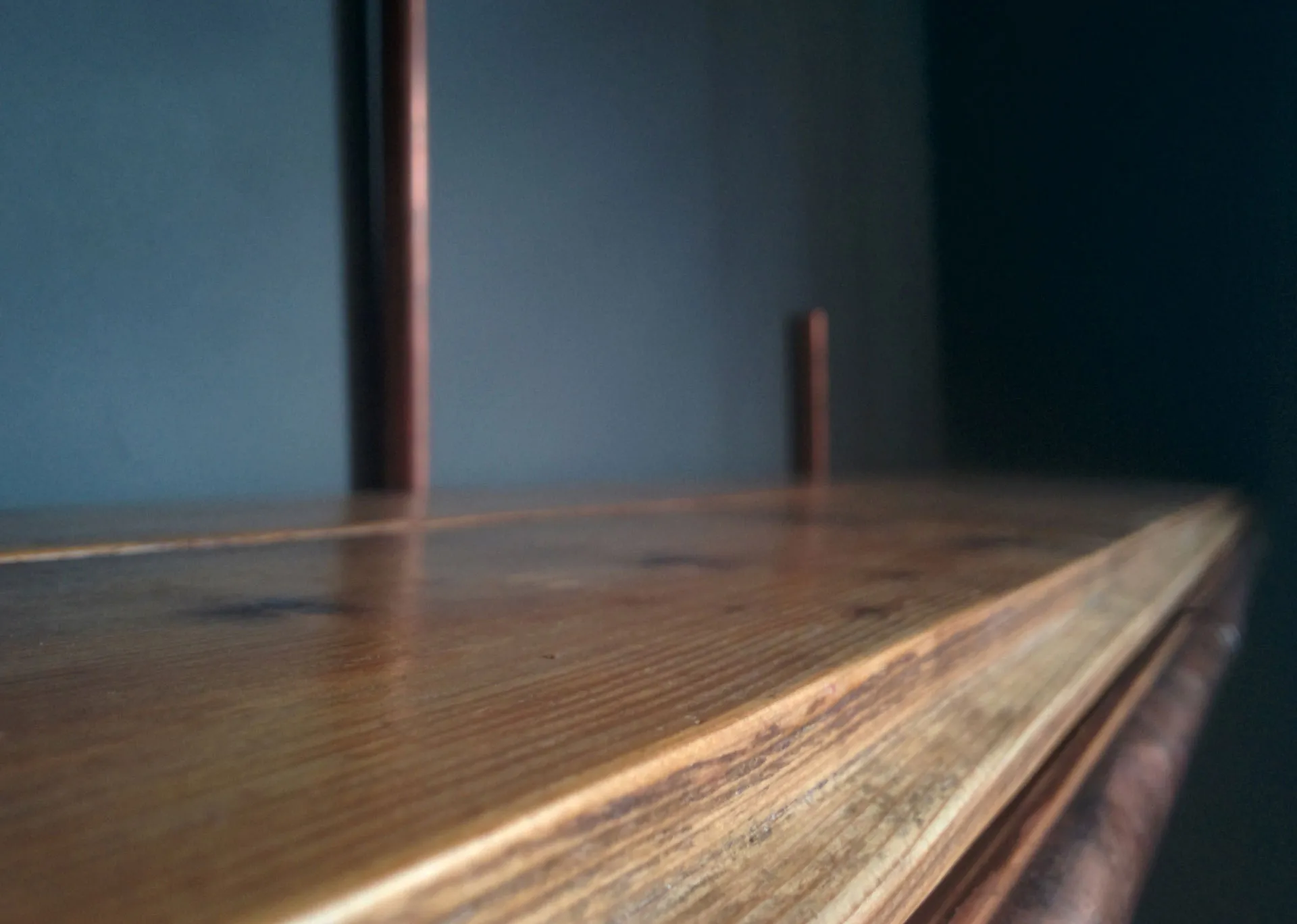 Low close-up along the oiled wooden shelf top, with two copper pipe uprights rising out of focus behind it.