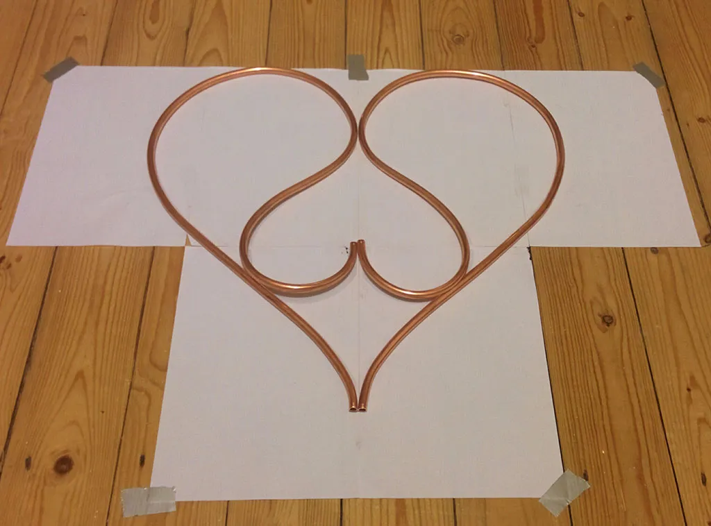 A double-heart shape of bent copper pipe laid over a paper template on a wooden floor.