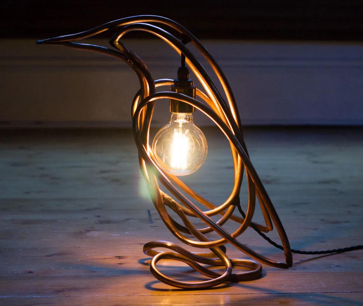 The lit penguin lamp on floorboards in low light, facing left, with the filament bulb glowing inside its body.