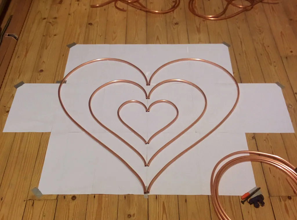 Three nested copper pipe hearts on a paper template, with a coil of unbent pipe beside them.