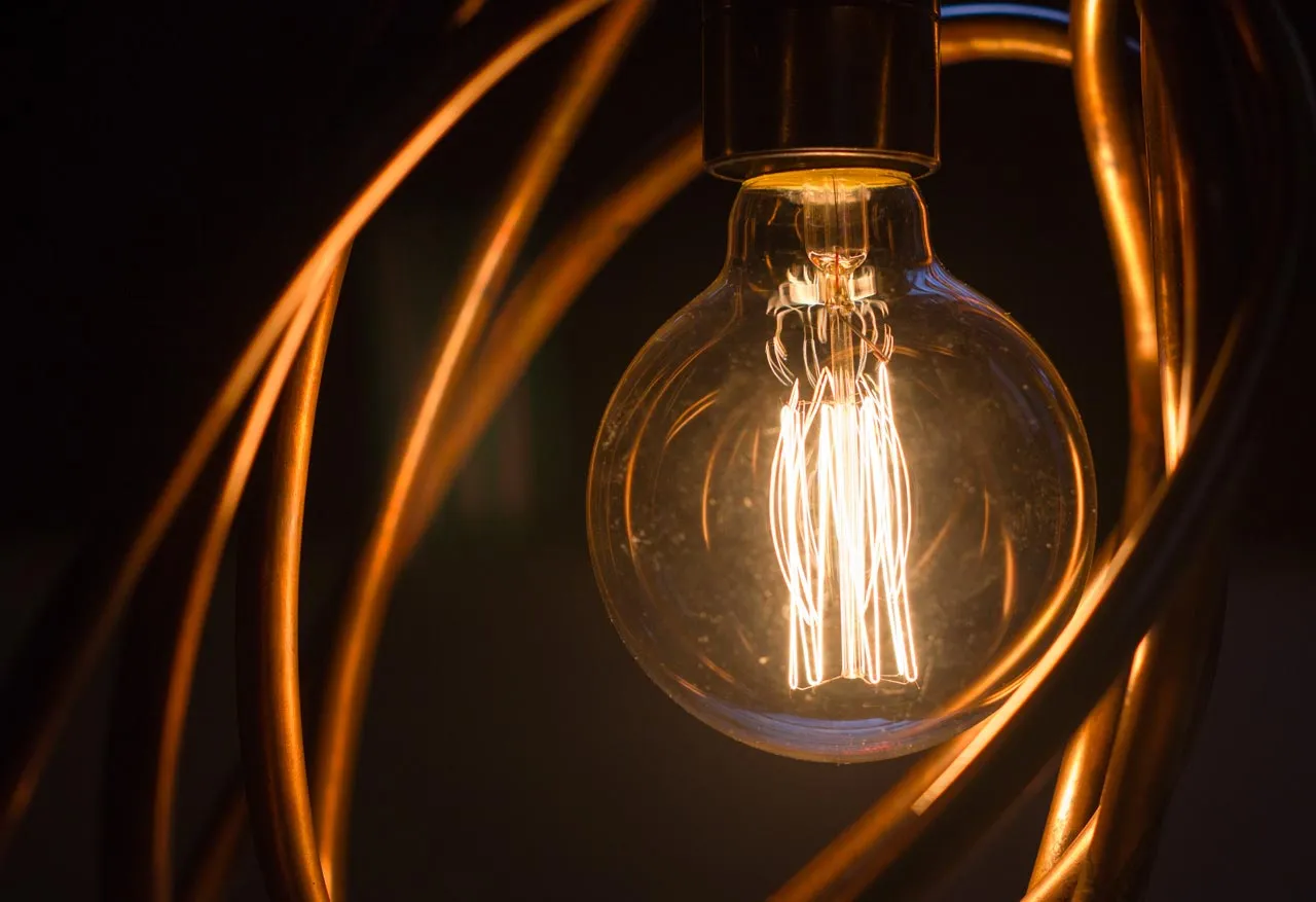 Close-up of the glowing filament bulb hanging from its dark lampholder, framed by curved copper pipes.