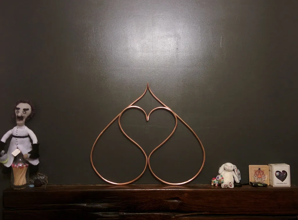 The double-heart standing on a wooden mantelpiece against a dark wall, between a knitted doll and some small ornaments.
