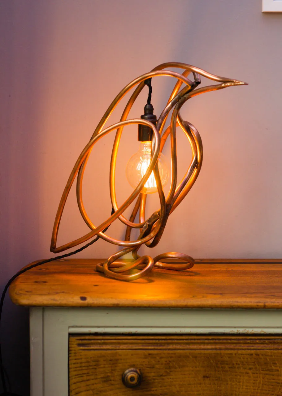 The copper pipe penguin lamp lit up on a wooden chest of drawers, facing right, with a filament bulb hanging inside its body.
