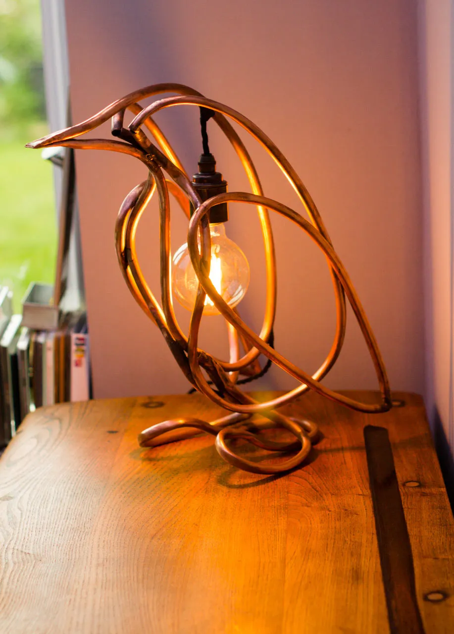 The lit penguin lamp facing left on a wooden table beside a window, with a row of books behind it.