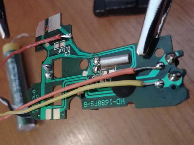 A small green clock circuit board with red and yellow wires soldered onto its contacts, held in a helping hand next to a battery.