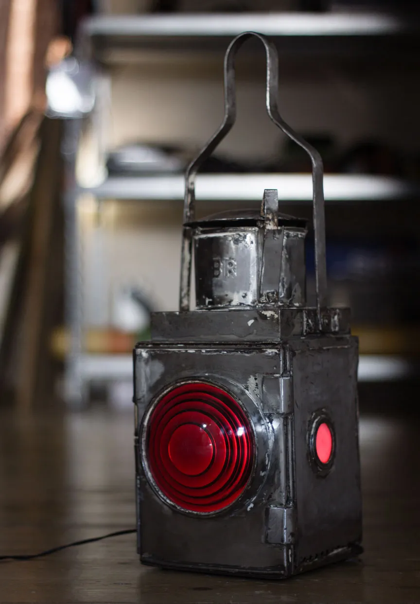 The restored lamp in bare, slightly paint-flecked metal on a floor, its red lens and side window lit, with BR stamped on the top.