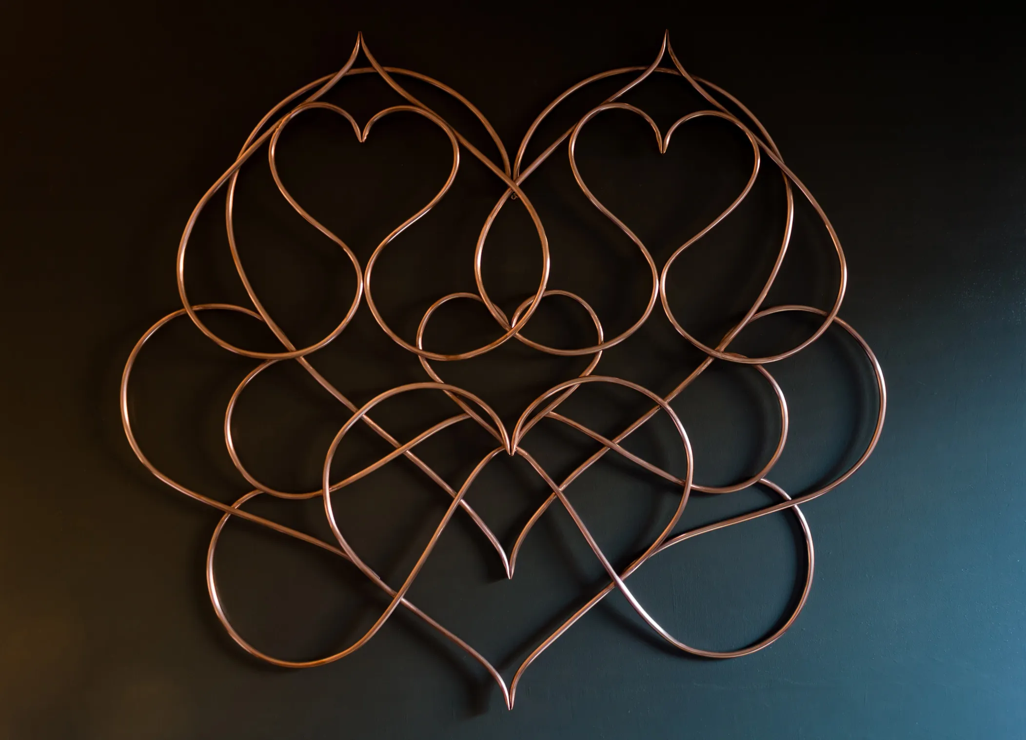 The finished copper pipe wall piece hanging on a dark wall, its interlocking heart curves catching the light.