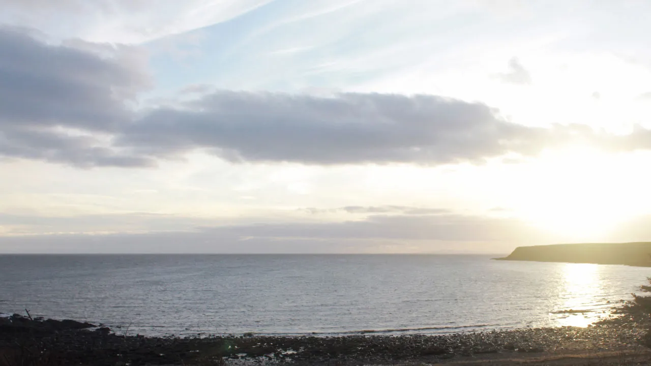 A photograph of a calm sea under a cloudy sky, with a low headland on the right lit by the setting sun and a pebbly shore in the foreground.